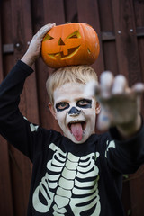 Happy young blond hair boy with skeleton costume holding jack o lantern. Halloween. Trick or treat. Outdoors portrait over wooden background