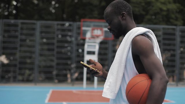 Young Muscly Serious Afro - American Guy in White Singlet Holding Ball and Using his Smartphone and Typing Message Walking at Street Basketbal Court. Technology and Sport Concept. Side View.