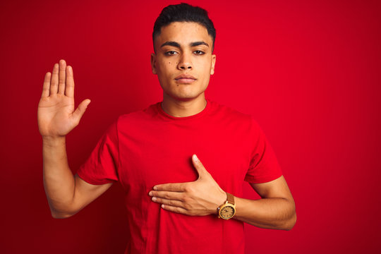 Young brazilian man wearing t-shirt standing over isolated red background Swearing with hand on chest and open palm, making a loyalty promise oath