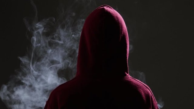 Young male wearing red hoodie vaping in a dark alley, exhales thick vapor