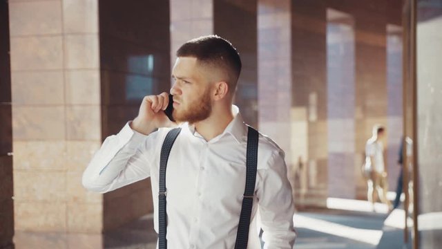 Close-up of brutal man with bristle walking out of the office and speaking by phone on the background of a beautiful modern building. Modern communication, dealing with problems, business talk. Male