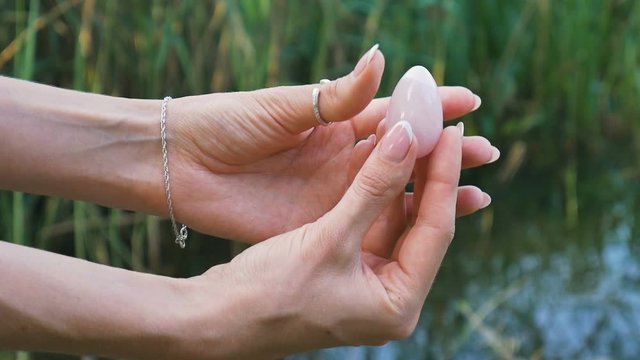 Female hand holding a rose quartz crystal yoni egg on river background. Women's health, unity with nature concepts
