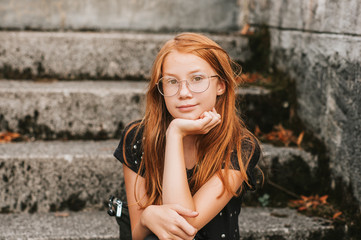 Outdoor portrait of sweet little girl with red hair, wearing eyeglasses