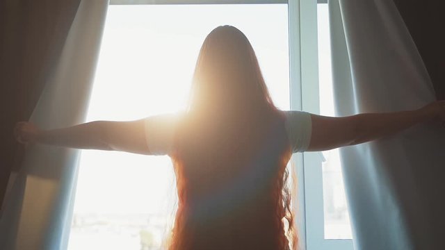 Slow motion with young woman opens dark curtains in morning and looks at morning sun light