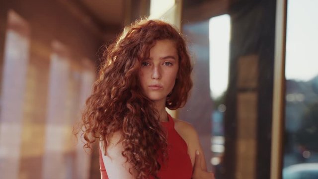 Close up view of extremely beautiful ginger curly student girl in attractive red dress proudly and disgruntledly posing on camera. Natural beauty, light makeup, outdoor shooting. Female portrait
