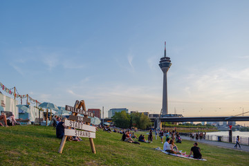 Outdoor sunny view of crowd of people sit, hang out and enjoy beautiful day on park at promenade riverside of Rhine River and background of Rhine Tower in Düsseldorf, Germany.   © Peeradontax