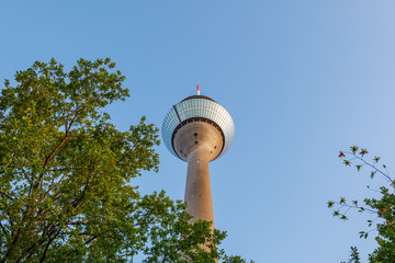 Low angle view of Rheinturm, high-rise radio or television tower with circular observation deck on the top over treetop against blue sky in Düsseldorf, Germany.