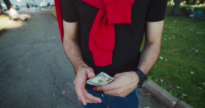 Portrait close-up young unsatisfied caucasian man making grimace showing small bundle of money salary standing oudoor in the park.
