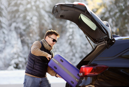 Handsome man going to vacations, loading his suitcase in car trunk