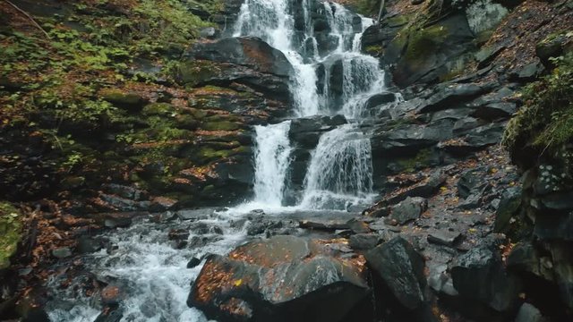 foaming water falls from grey wet rocks on high hill surrounded by green grass and fallen leaves aerial view. Shipot waterfall, Carpathian mountains. 4K