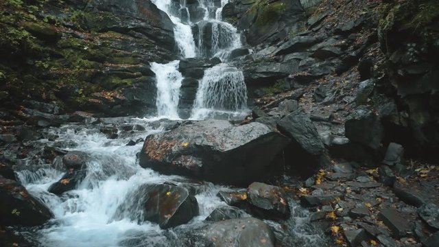 foaming mountain waterfall water surrounded by grey wet stones with fallen leaves and moss upper view. Shipot waterfall, Carpathian mountains. 4K