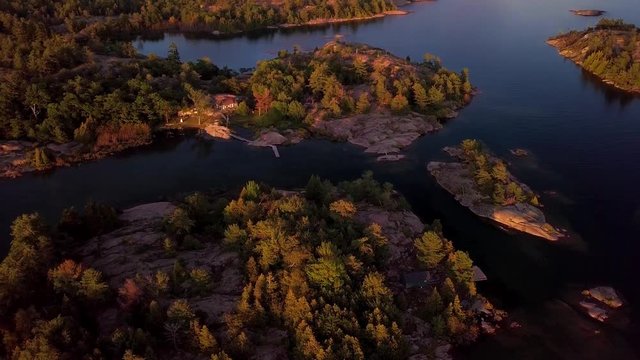 Abandoned Cabin on Rocky Island with Pine Trees in Blue Lake at Sunset, Drone Aerial Wide Pedestal Up Tilt Down Overhead. Fly over Natural Georgian Bay Shore at Summer Golden Hour, Ontario Canada