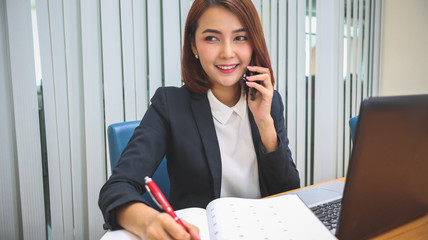 Happy smiling Business woman sitting at table and taking notes in laptop.On table is laptop while talking on phone