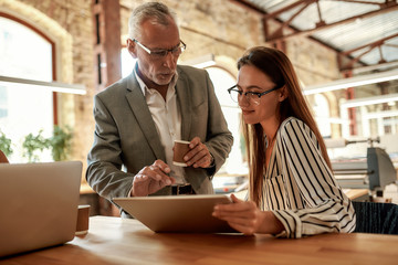 Senior man in formal wear holding coffee cup and explaining something to his young female colleague while working together in the modern office