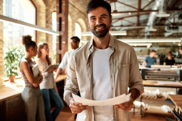 Great working day! Young bearded man in casual clothes holding documents and looking at camera with smile while standing in the modern office