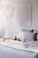 Birthday breakfast on white wooden trey on large bed with grey sheets and blanket, bunch of white balloons above nightstand, copy space on the empty wall