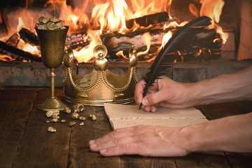 Gold nuggets in a goblet, golden crown and royal decree or order document on the wooden table over burning fire background.