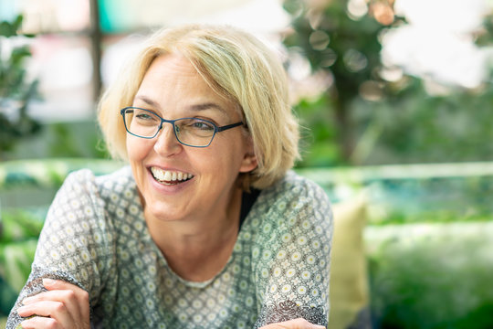 Closeup outdoor nature portrait of blond happy smiling mature woman in her fifties wearing glasses.