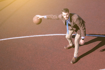 Stylish bearded handsome hipster guy in a trendy suit in a jacket and tie on a basketball court playing basketball. Fashionable handsome man with a ball in his hand, top view. Business concept, sport.