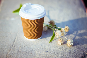Paper cup with coffee. Dry flowers on brown background. Bright summer good morning. A bee flies near a coffee cup. Brown paper cup with plastic white lid.