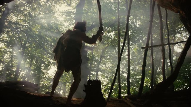 Primeval Caveman Wearing Animal Skin Holds Stone Tipped Spear, Stands at the Cave Entrance Looking over Prehistoric Forest Ready to Hunt Animal Preys. Neanderthal Going Hunting in the Jungle