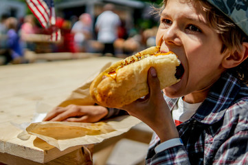 Boy eating barbecue grilled hot dog on family picnic celebrating independence day with flag on the background