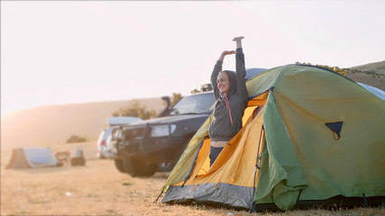 Young woman wakes up and comes out from tourist tent and stretches at campsite in autumn windy weather. Expedition camp with off-road vehicle on the background.