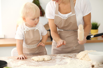 Little girl and her blonde mom in beige aprons playing and laughing while kneading the dough in kitchen. Homemade pastry for bread, pizza or bake cookies. Family fun and cooking concept