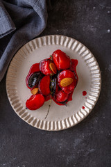 red ripe caramelised plums with almonds on white plate on dark background. top view