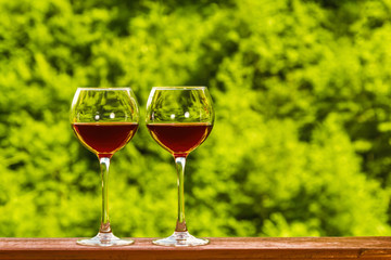 Two glasses of red wine on the deck of a wooden house on a green fresh forest background. Copyspace.