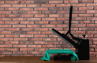 Heat press machine with t-shirt on wooden table near brick wall, space for text