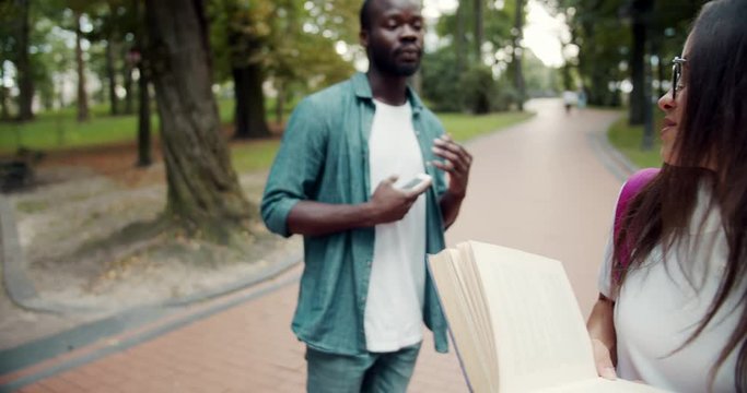 Afro-american male with smartphone hitting young female student in eyeglasses reading book during walk