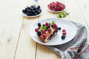 Piece of pie with blueberries, rasberry and mint for dessert on a white plate, napkin. Pieces of delicious homemade cake on a wooden boards background