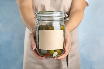 Woman holding jar of pickled cucumbers with blank sticker against blue background, closeup view. Space for text