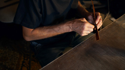 Tanner works with needle and thread. Man sews leather details clamped in vice. Dark workshop, copy space.