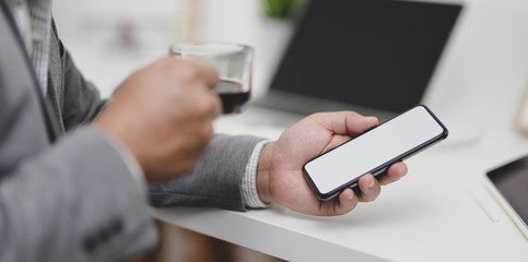 Close-up view of businessman holding smartphone while drinking a cup of coffee in his modern office