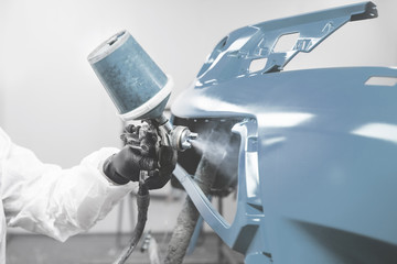 Male worker in protective clothes and mask painting car bumper using spray paint.