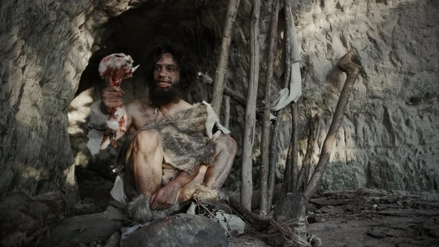 Primeval Caveman Wearing Animal Skin Holds Bone and Hits Rock with It. Neanderthal Fooling Around near the Cave Entrance, Maybe Creating first Primitive Tools or Weapons by Accident.
