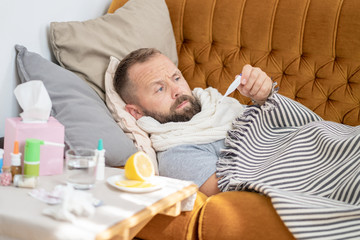 Sick man lying on sofa checking his temperature at home in the living room. Man in scarf hold thermometer close up. Measure temperature. High temperature concept. Break fever remedies.