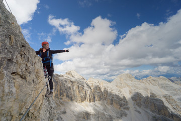 attractive brunette female climber on a steep and exposed Via Ferrata in the Dolomites pointing to the distance