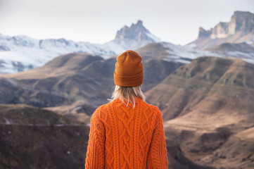 Portrait from the back of the girl traveler in an orange sweater and hat in the mountains against...