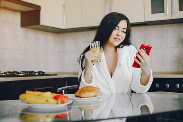 beautiful young girl with black hair and white robe sitting at home in the kitchen at the table and eating an croissant and drinking orange juice