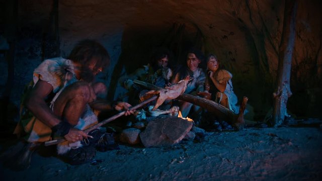 
Neanderthal or Homo Sapiens Family Cooking Animal Meat over Bonfire and then Eating it. Tribe of Prehistoric Hunter-Gatherers Wearing Animal Skins Eating in a Dark Scary Cave at Night. Zoom in Shot