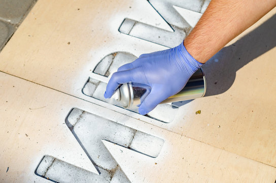 Hand holds an aerosol spray can, puts the image on the asphalt through a stencil