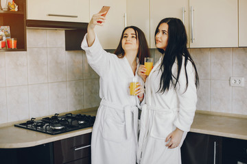 two beautiful young girls with black hair and white robe standing at home in the kitchen and drinking a orange juice