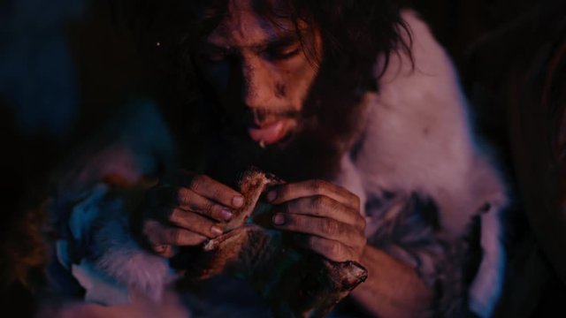 
Portrait of Tribe Leader Wearing Animal Skin Eating in a Dark Scary Cave at Night. Neanderthal or Homo Sapiens Family Cooking Animal Meat over Bonfire and then Eating it. Close-up Shot