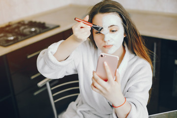 A young girl in a white dressing gown with hands hands sitting in the kitchen and applying a refreshing mask with a brush on her face