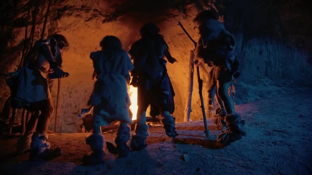 Tribe of Prehistoric Hunter-Gatherers Wearing Animal Skins Dance Around Bonfire Outside of Cave at Night. Neanderthal / Homo Sapiens Family Doing Pagan Religion Dancing Near Fire Back View Slow Motion