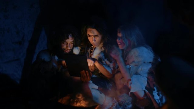 Tribe of Prehistoric, Primitive Hunter-Gatherers Wearing Animal Skins Use Digital Tablet Computer in a Cave at Night. Neanderthal or Homo Sapiens Family Browsing Internet, Watching Videos, Streaming