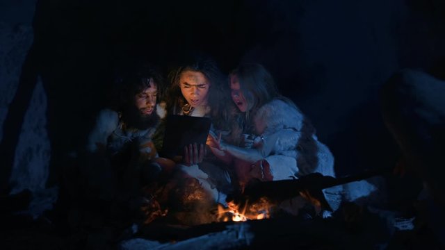 Tribe of Prehistoric, Primitive Hunter-Gatherers Wearing Animal Skins Use Digital Tablet Computer in a Cave at Night. Neanderthal or Homo Sapiens Family Browsing Internet, Watching Videos, Streaming